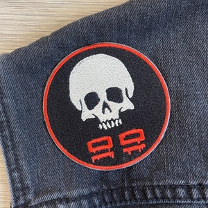 May include: A black and red embroidered patch featuring a white skull and crossbones design. The skull has a white face with black eyes and a black mouth. The crossbones are red and white. The patch is sewn onto a black denim jacket.