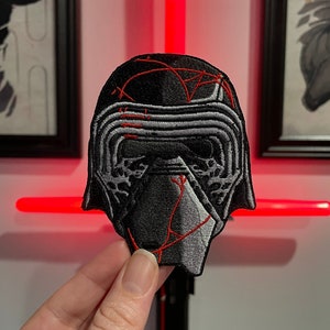 May include: A black and gray embroidered patch of Kylo Ren's helmet from Star Wars. The helmet has red stitching details.