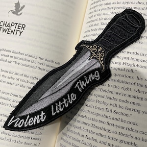 May include: A black and white embroidered patch of a dagger with the words "Violent Little Thing" on it. The patch is on a book with the text "Chapter Twenty" visible.