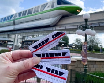 May include: Three white embroidered patches of the Disney monorail, each with a red stripe and black details. The patches are held in a hand in front of a monorail track with a green and white monorail train.