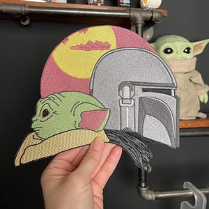 May include: Embroidered patch featuring a green alien character with large ears, wearing a Mandalorian helmet, against a yellow and red background with a cloud.