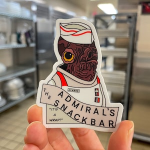 May include: A white sticker with a cartoon illustration of a brown alien wearing a white uniform and a red and white paper hat. The alien is holding a banner that says "The Admiral's Snack Bar" with the text "Ackbar" in a smaller font above the banner. The text "It's a Wrap!" is below the banner.