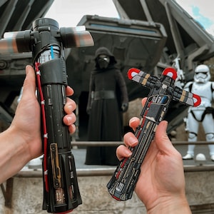 May include: Two Star Wars lightsaber replicas, one red and one black, are held in the hands of two people. The lightsabers are detailed with intricate designs and have a metallic finish. The background features a Star Wars themed backdrop.