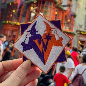 May include: A white, purple, and orange embroidered patch with two witches, one in purple and one in orange, holding wands. The patch is shaped like a star and has a white background.