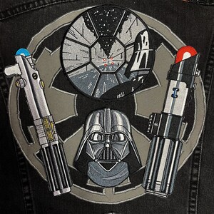 May include: A black denim jacket with a gray embroidered Star Wars symbol. The symbol features a Death Star, Darth Vader, a lightsaber, and a light saber.