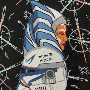 May include: Embroidered patch of Ahsoka Tano from Star Wars. She is wearing her blue and white Togruta headtails and her white and blue clone trooper helmet. The helmet has a blue visor and a black eye piece. The patch has a white background and is surrounded by a black border.