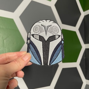 May include: A white and blue embroidered patch of a Mandalorian helmet. The helmet has a white visor and a black T-shaped design on the front.