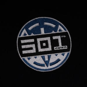 May include: A white and blue embroidered patch with the text "301" in white and a small square in the center. The patch is circular and has a geometric design around the edge.