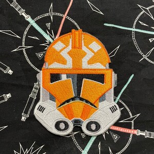 May include: An orange and white embroidered patch of a Star Wars clone trooper helmet. The helmet has a white visor and a black stripe across the forehead.