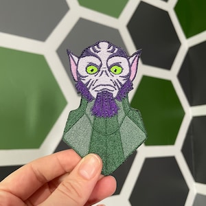 Könnte beinhalten: Ein gestickter Aufnäher mit einem grünen, violetten und weißen Außerirdischen mit großen Augen und einem violetten Bart. Der Außerirdische trägt einen grünen, rautenförmigen Kragen.