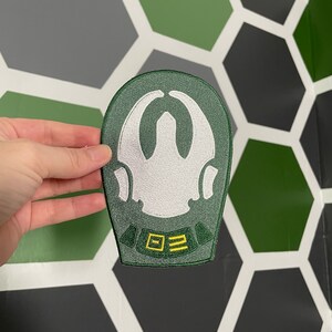 Könnte beinhalten: Grüner gestickter Aufnäher mit einem weißen Symbol und gelbem Text, der "0Ξ2" lautet.