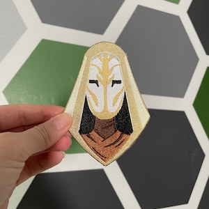 May include: Embroidered patch featuring a stylized figure wearing a white mask and a brown hooded robe.