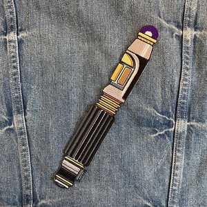 May include: A black and gold embroidered patch of a sonic screwdriver, a fictional tool used by the Doctor in the British science fiction television series Doctor Who.