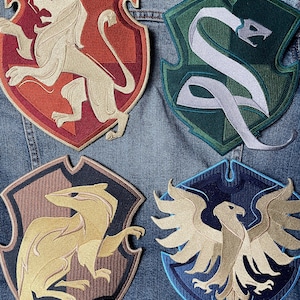 May include: Four embroidered patches featuring the house crests from the Harry Potter series. The crests are on a blue denim background. The crests are Gryffindor (lion), Slytherin (snake), Hufflepuff (badger), and Ravenclaw (eagle).