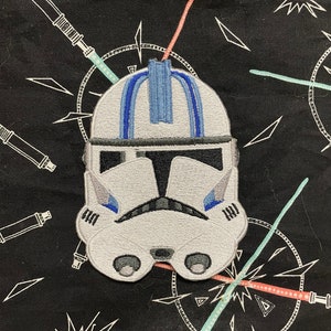 May include: A white and blue embroidered patch of a Star Wars clone trooper helmet. The helmet has a blue stripe down the center and a black visor.