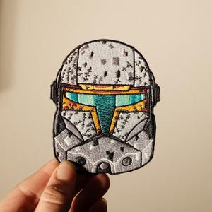 May include: Embroidered patch featuring a detailed helmet design with a gray, speckled pattern. The helmet has a visor with teal, yellow, and orange accents. The patch is held by a hand.