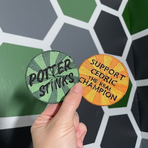 Peut inclure: Deux patchs ronds brodés. L'un est vert avec le texte "Potter Stinks" en noir. L'autre est orange avec le texte "Support Cedric The Real Champion" en noir.