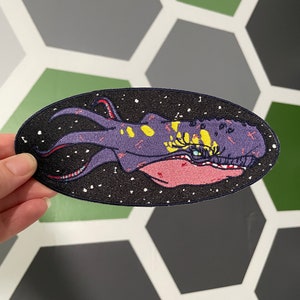 Könnte beinhalten: Ein gestickter Aufnäher mit einer violetten und gelben Weltraumkreatur mit einem großen Auge und Tentakeln. Die Kreatur ist vor einem schwarzen Hintergrund mit weißen Sternen platziert.