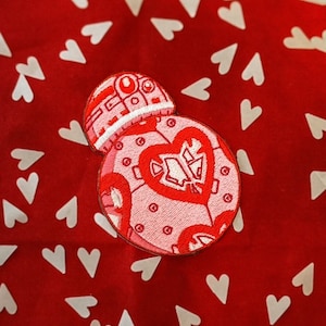 Puede incluir: Un parche bordado rosa y blanco de un personaje robot con un diseño de corazón sobre un fondo rojo con corazones blancos.