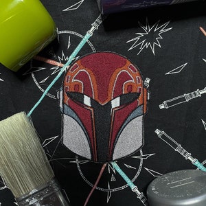 May include: A red and white embroidered patch of a Mandalorian helmet with a black background.