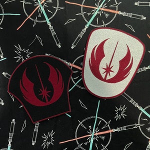 May include: Two embroidered patches featuring a stylized emblem with a starburst design. One patch is black with burgundy details, the other is white with burgundy details. The background is black with a repeating pattern of lightsabers.
