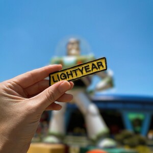 May include: A yellow and black patch with the word "LIGHTYEAR" in black lettering. The patch is being held in a hand with a blurred image of a large, white, space ranger figure in the background.
