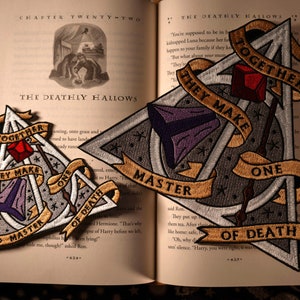 May include: Two embroidered patches featuring the Deathly Hallows symbol from the Harry Potter series. The larger patch is gold, white, and gray with the text "Together they make one Master of Death". The smaller patch is similar but smaller and has a red, purple, and white color scheme. The text on the smaller patch reads "Together they make one Master of Death".
