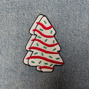 May include: Embroidered patch in the shape of a Christmas tree. The tree is white with red wavy lines and green sprinkles. The patch has a black border and is set against a blue denim background.