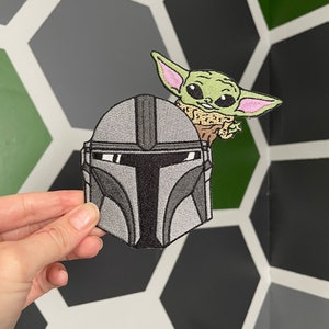 May include: Embroidered iron-on patch featuring a gray Mandalorian helmet with a green Baby Yoda character peeking out from behind it.