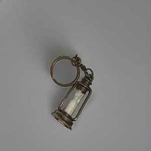 May include: A metal keychain featuring a miniature lantern-shaped charm. The charm has a dark, weathered bronze-tone finish and contains a small, white, hourglass-shaped insert within its frame. It is attached to a standard circular metal ring.