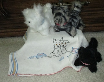 May include: Three plush toy dogs, one white, one black, and one gray and white, are sitting on a white embroidered tea towel with a blue and yellow border. The towel has an embroidered design of three dogs sitting around a table with teacups.