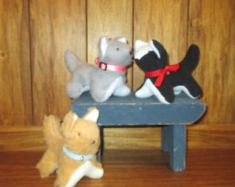 May include: Three plush toy dogs, one gray, one black and white, and one brown, are sitting on a small blue wooden stool. The gray and black and white dogs are facing each other, while the brown dog is facing the camera.