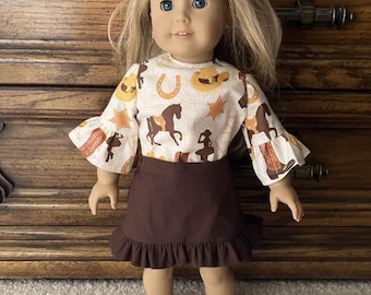 May include: A doll wearing a brown skirt with a ruffle hem and a white long-sleeved shirt with a western print featuring horseshoes, stars, and cowboy hats.