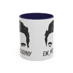 May include: A white ceramic mug with a dark blue interior and rim. The exterior features a repeating black graphic illustration of a person's face and the text "EW, MISOGYNY" printed in a bold, sans-serif font on the white surface.