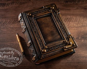 May include: A dark brown leather-bound journal with a rectangular centre panel, intricate embossed scrollwork borders, and metallic corner accents. The spine features a diamond-patterned texture and metal studs. A gold-toned pen rests beside the journal.