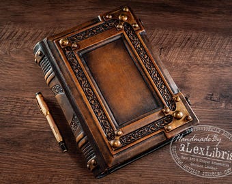 Könnte beinhalten: Ein braunes, in Leder gebundenes Notizbuch mit geprägter Blumenbordüre, messingfarbenen Metallecken und einem rechteckigen Mittelfeld. Ein goldfarbener Stift liegt neben dem Buch, das den Text Handmade By aLexLibris aufweist.