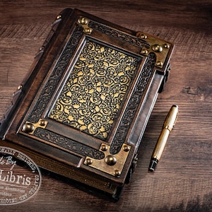May include: A handcrafted, antique-style journal with a detailed, ornate cover. The journal features a dark brown leather exterior with brass corner accents and a gold patterned center panel. A gold pen rests beside the journal on a wooden surface.