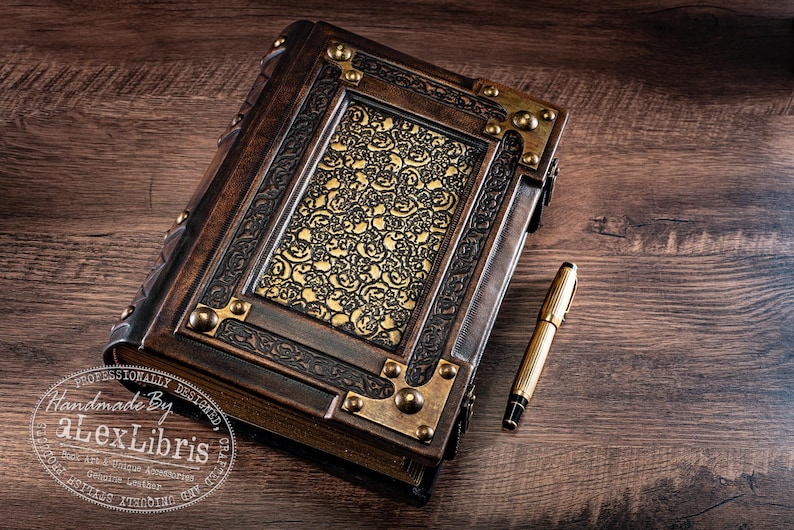 May include: A handcrafted, antique-style journal with a detailed, ornate cover. The journal features a dark brown leather exterior with brass corner accents and a gold patterned center panel. A gold pen rests beside the journal on a wooden surface.