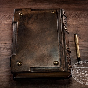 May include: A handcrafted leather-bound journal with brass accents and a matching gold pen. The journal is a rich brown color, with a textured surface and brass rivets. The pen is gold with black accents. The journal is on a wooden surface.
