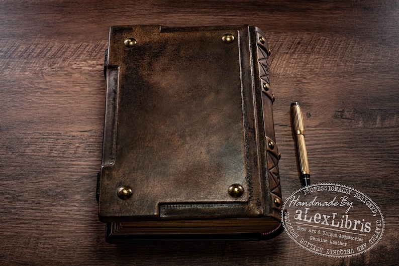 May include: A handcrafted leather-bound journal with brass accents and a matching gold pen. The journal is a rich brown color, with a textured surface and brass rivets. The pen is gold with black accents. The journal is on a wooden surface.