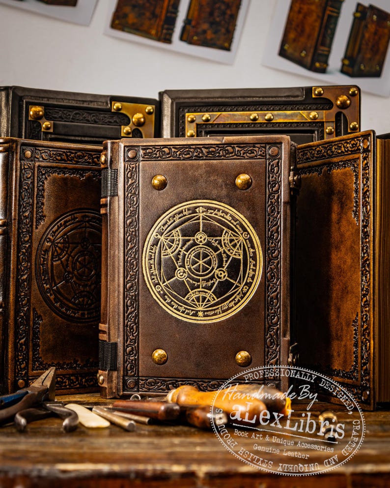K&ouml;nnte beinhalten: Eine Sammlung handgefertigter, ledergebundener B&uuml;cher mit aufwendigen gepr&auml;gten Designs. Die braunen Einb&auml;nde sind mit goldenen Akzenten versehen, darunter dekorative Nieten und kreisf&ouml;rmige Symbole. Die B&uuml;cher werden mit Werkzeugen pr&auml;sentiert, was auf Handarbeit hindeutet.