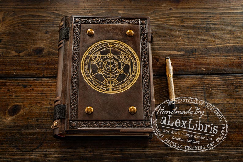 K&ouml;nnte beinhalten: Ein braunes Lederbuch mit einem goldenen, kreisf&ouml;rmigen Design auf dem Einband und goldenen Nieten. Ein goldener Stift liegt neben dem Buch. Ein handgefertigtes Produkt.