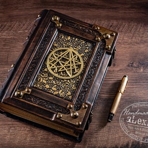 May include: A handcrafted, leather-bound journal with brass accents and a gold emblem. A gold pen rests beside the book on a wooden surface. The journal features intricate detailing and a vintage aesthetic.
