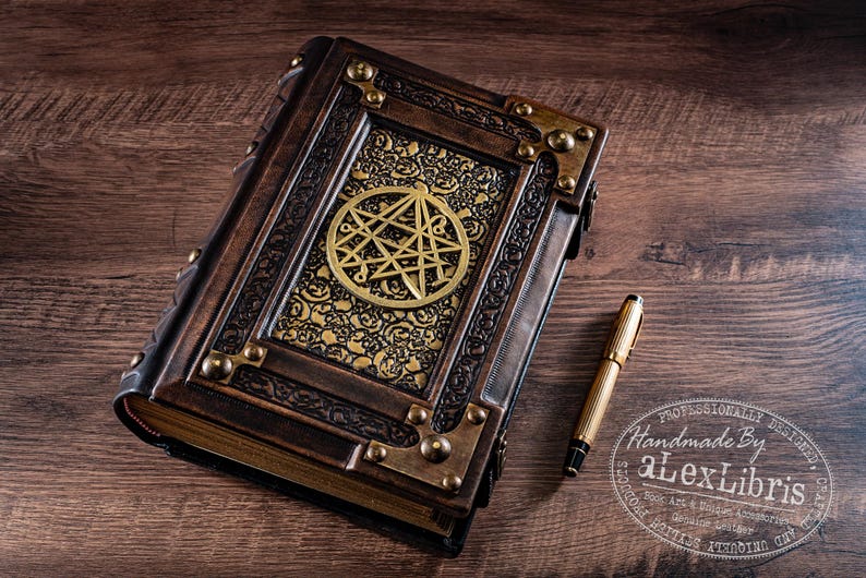 May include: A handcrafted, leather-bound journal with brass accents and a gold emblem. A gold pen rests beside the book on a wooden surface. The journal features intricate detailing and a vintage aesthetic.