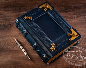 May include: A dark blue leather-bound journal featuring ornate gold-toned metal corner protectors, decorative studs, and an embossed border pattern. A white and black patterned pen is positioned next to the journal, which displays the text aLexLibris.