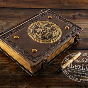 K&ouml;nnte beinhalten: Ein braunes, ledergebundenes Buch mit goldenen Akzenten und einem kreisf&ouml;rmigen Emblem auf dem Einband. Das Buch hat goldfarbene Seiten und einen goldenen Stift. Der Text "Handmade By Lex Libris" ist sichtbar. Es ist wahrscheinlich ein Tagebuch.