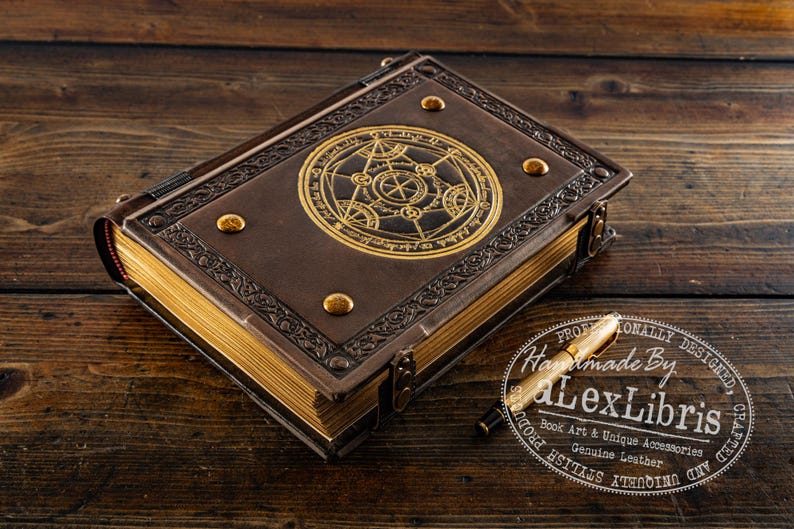 K&ouml;nnte beinhalten: Ein braunes, ledergebundenes Buch mit goldenen Akzenten und einem kreisf&ouml;rmigen Emblem auf dem Einband. Das Buch hat goldfarbene Seiten und einen goldenen Stift. Der Text "Handmade By Lex Libris" ist sichtbar. Es ist wahrscheinlich ein Tagebuch.