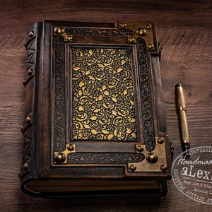 May include: A handcrafted leather-bound journal with ornate gold accents and a decorative floral design on the cover. The journal is dark brown with brass corner details and a gold pen resting beside it. The text "Handmade by alexLibris" is visible.