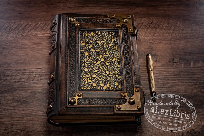 May include: A handcrafted leather-bound journal with ornate gold accents and a decorative floral design on the cover. The journal is dark brown with brass corner details and a gold pen resting beside it. The text "Handmade by alexLibris" is visible.