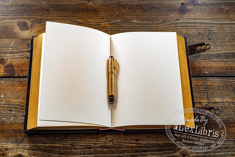 Op de afbeelding: Een open boek met blanco pagina's en een gouden pen in het midden. Het boek heeft een donkerbruine leren kaft en goudkleurige randen. De achtergrond is een houten oppervlak.